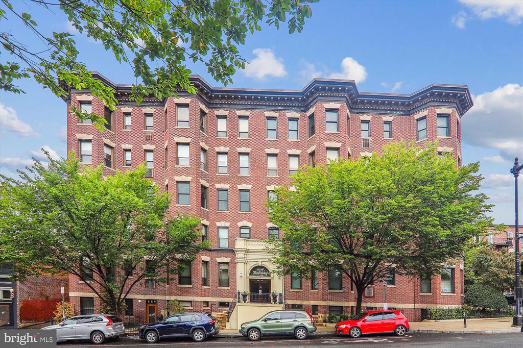 Red-brick apartment building with a decorative roofline, central entrance, and leafy trees along the street.
