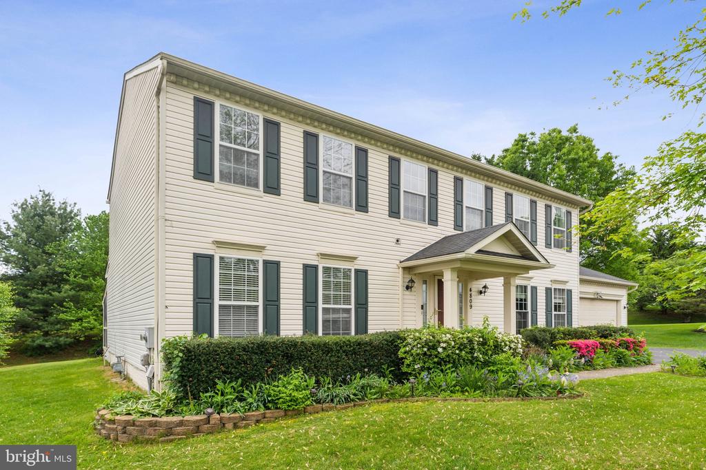 Two-story beige house with green shutters, a covered entrance, and landscaped garden beds.