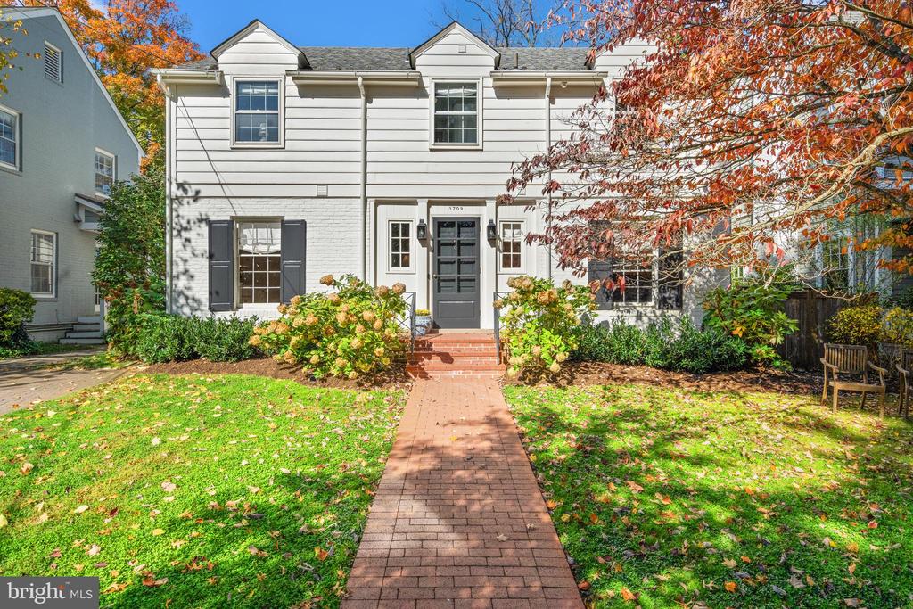 White two-story house with dark shutters, brick walkway, and autumn foliage.