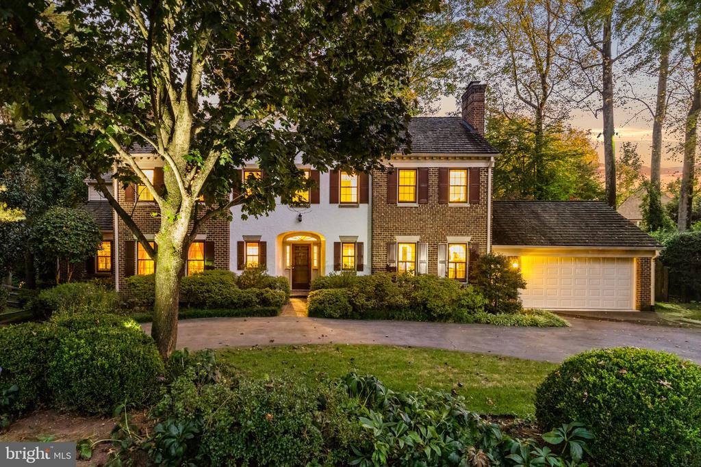 Two-story brick house with an attached garage, illuminated windows, and mature trees at dusk.