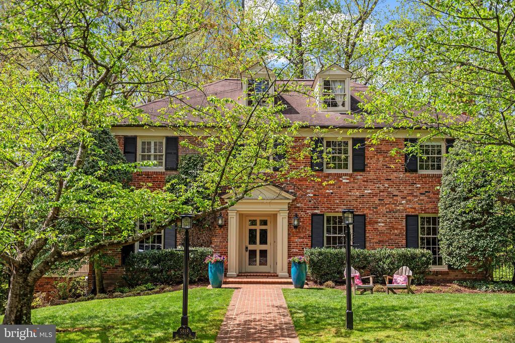 Red brick house framed by leafy trees, with a brick walkway, pale entry, and dormer windows.