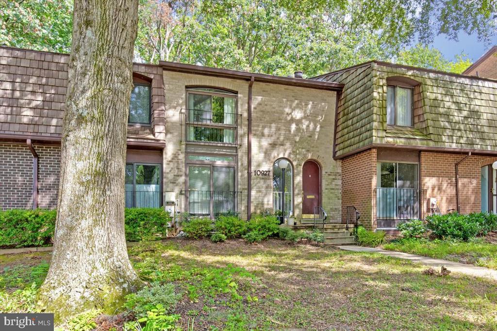 Multi-level brick and shingle residence with a burgundy arched door, balconies, and a large foreground tree.