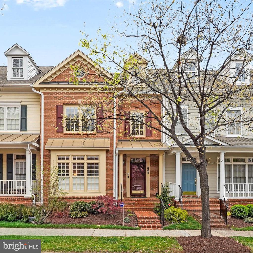 Red-brick townhouse with burgundy shutters and door, brick steps, and a budding tree in front.