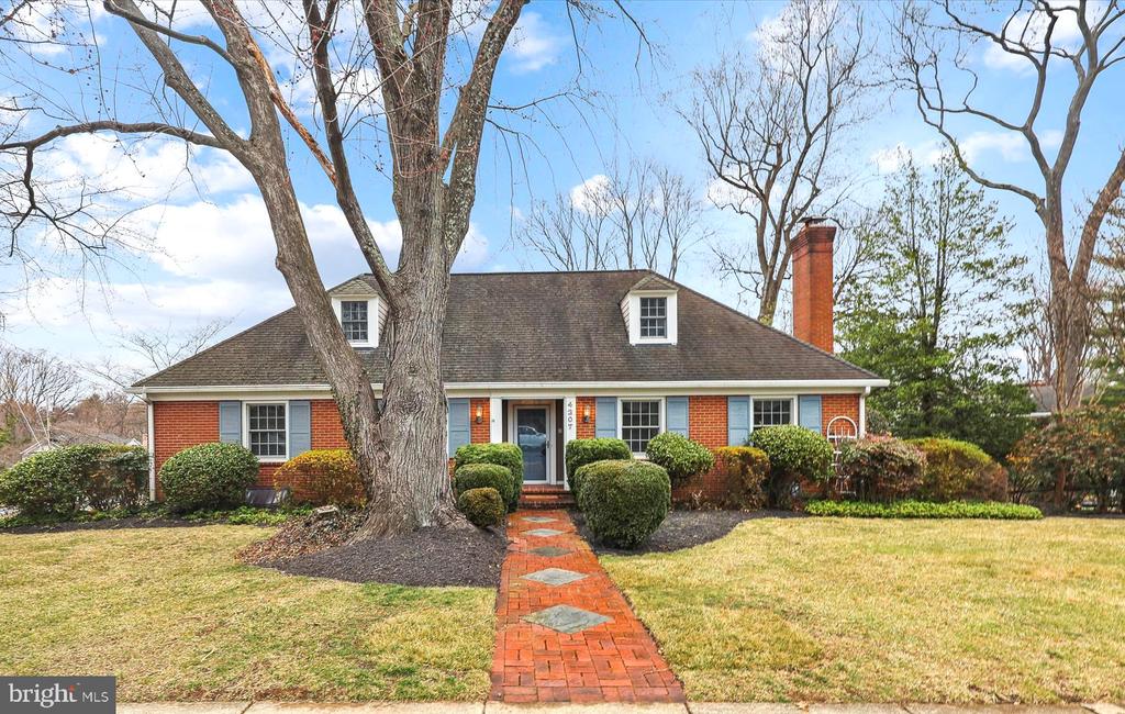 Red brick house with blue shutters, dormer windows, and a brick walkway beneath mature trees.
