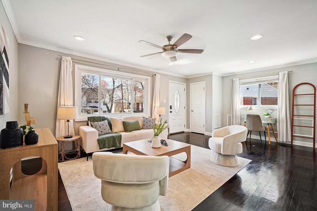 Staged living room with a cream sofa, large front window, ceiling fan, and dark hardwood floors.