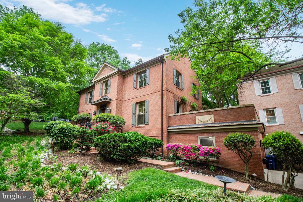 Two-story brick house with pale shutters, a low brick wall, and lush landscaped gardens.