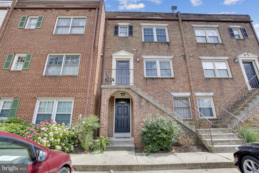 Brick townhome exterior with raised entry stairs, blue doors, flowering shrubs, and cars along the curb.