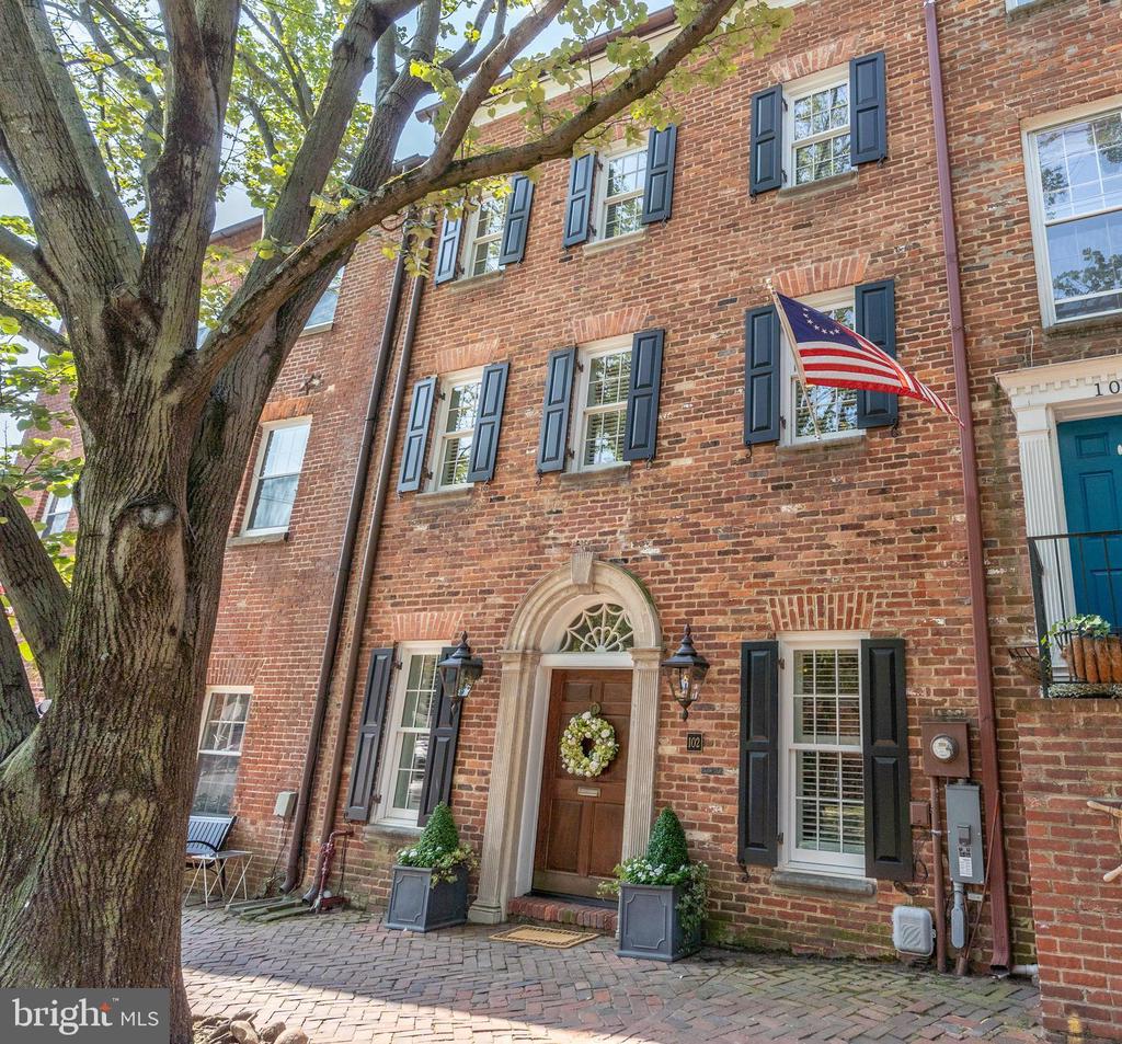 Red brick townhouse with dark shutters, an arched entrance, and an American flag beneath a leafy tree.