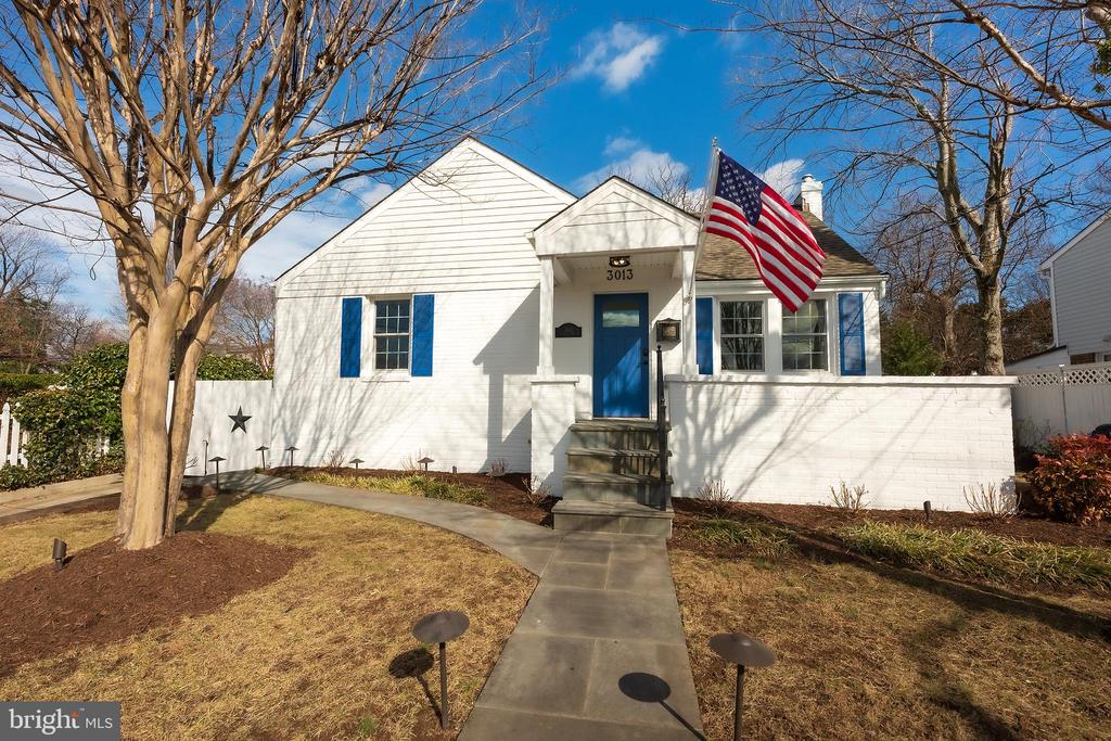 White house with blue shutters and front door, a U.S. flag, and a walkway through the front yard.