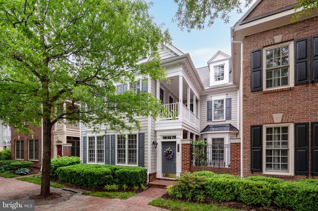 Townhouse exterior with a white balcony, brick facade, dark shutters, and leafy street tree.