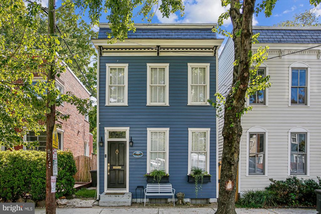 Blue two-story house with white trim, window boxes, and a bench beneath a large street tree.