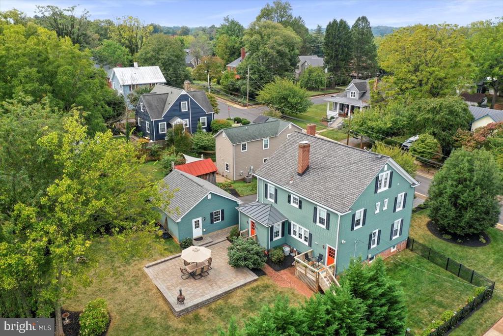 Aerial view of a green home with a fenced yard and paved patio, surrounded by leafy trees and neighboring houses.