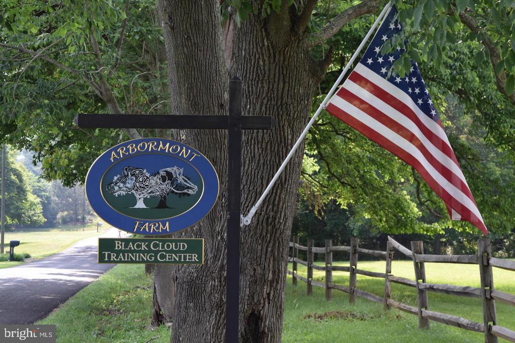 Arbormont Farm entrance sign beside a tree, American flag, and wooden fence.