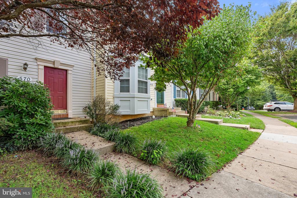 Attached home with a red entry door, landscaped garden, mature trees, and sidewalk