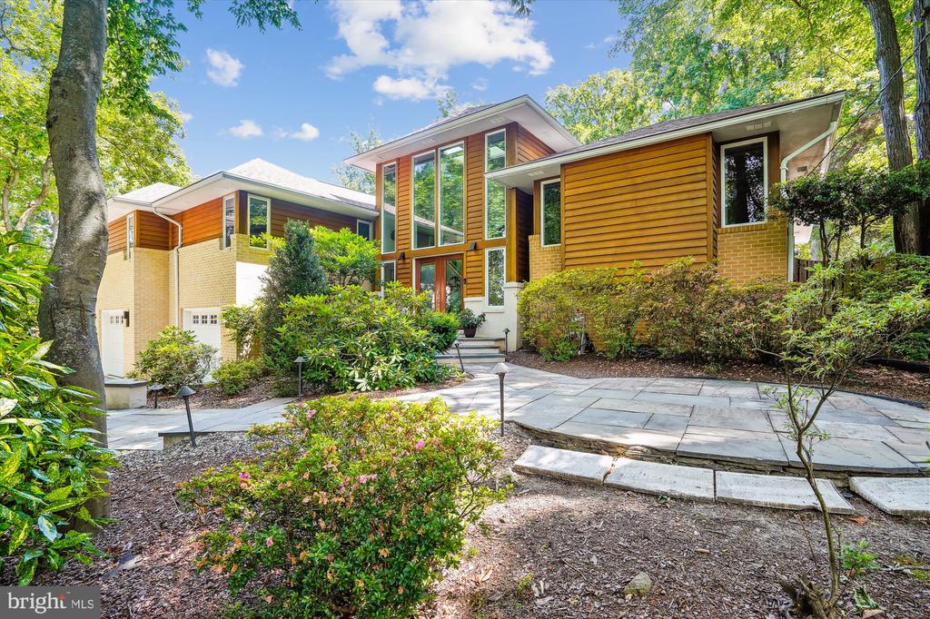 Contemporary wood-and-brick house with large windows, a paved approach, and mature trees.