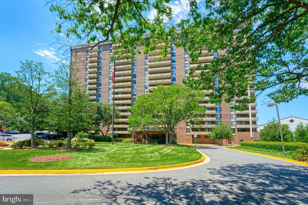 Brick condominium building with balconies, framed by leafy trees above a landscaped driveway.