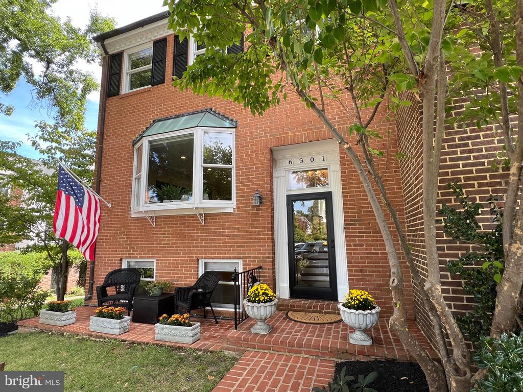 Red-brick home with a bay window, brick patio seating, flower planters, and an American flag.