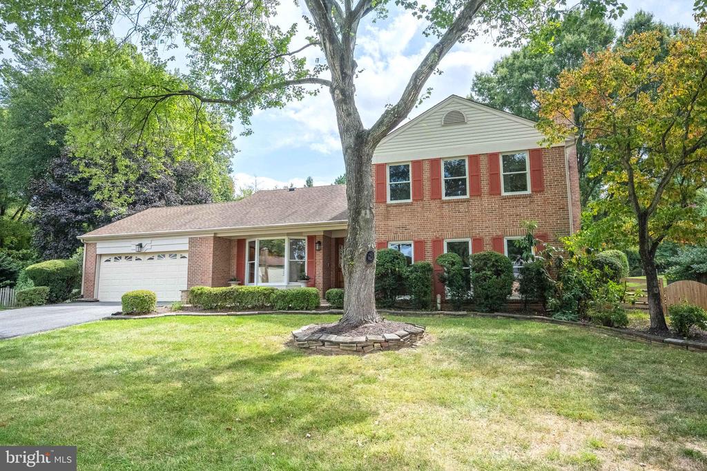 Two-story brick house with red shutters, an attached garage, and a large tree in the front lawn.