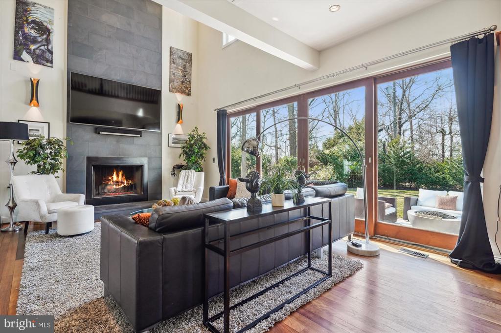 Living room with a gray tile fireplace wall, dark leather sectional, and large windows overlooking trees.