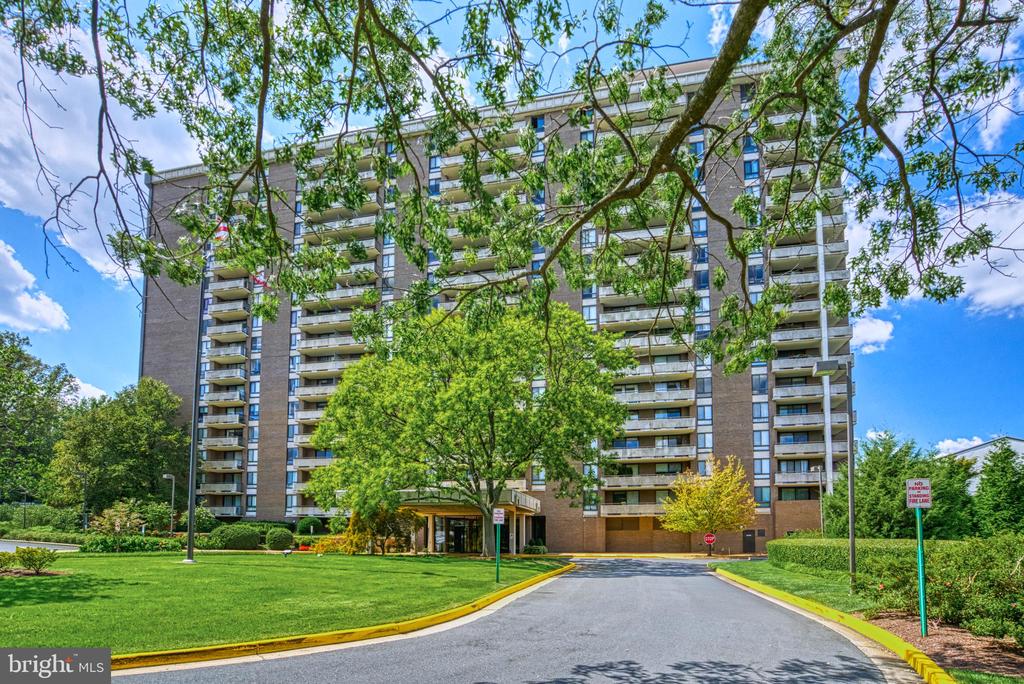 Condominium building with rows of balconies, leafy trees, landscaped grounds, and a front driveway.