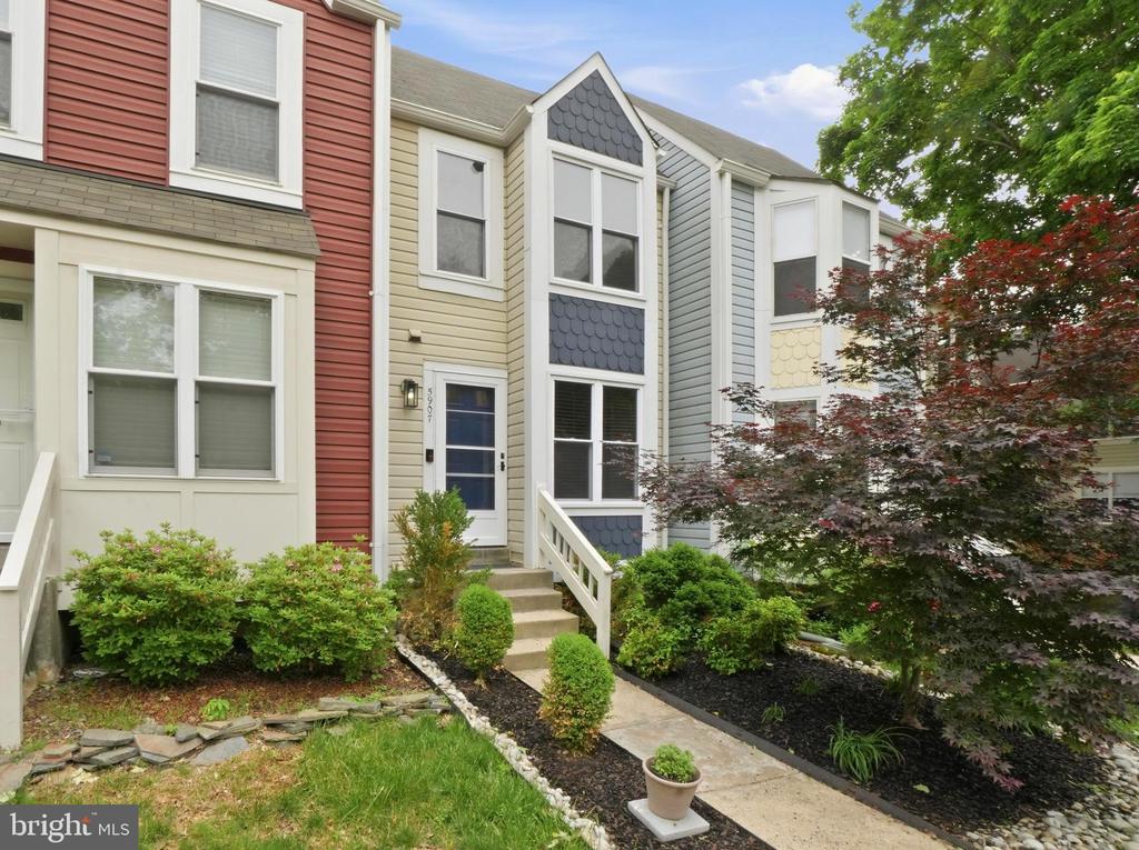 Beige townhome with a blue-gray decorative gable, front steps, and landscaped shrubs.
