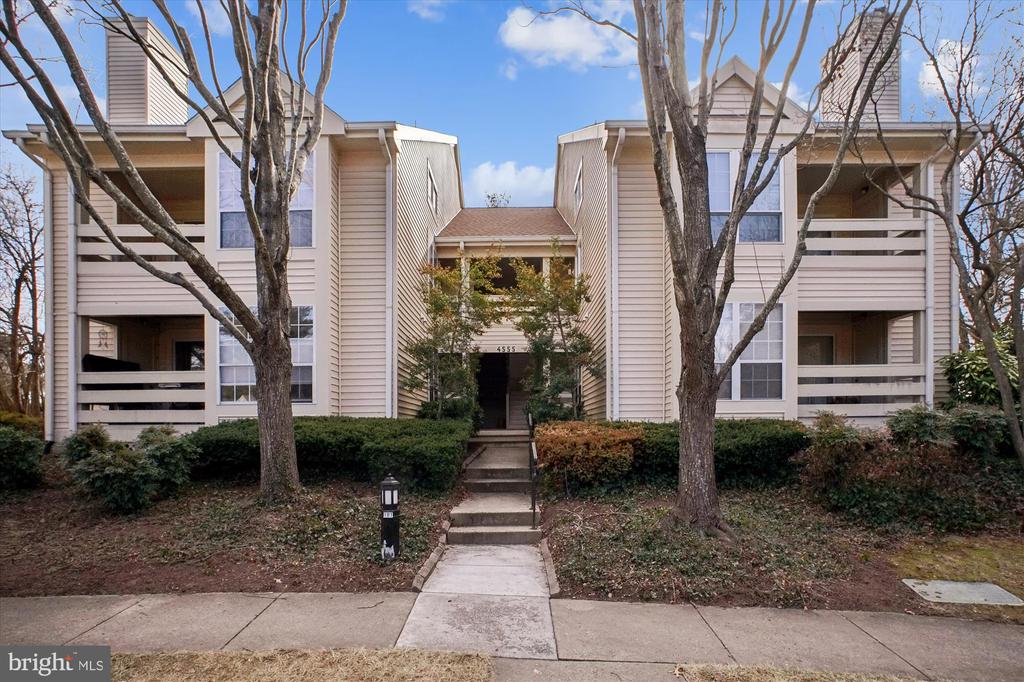 Two-story beige condominium building with balconies and a central entrance framed by leafless trees.