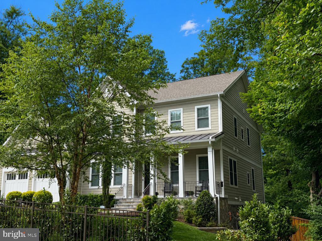 Two-story beige house with a covered porch, attached garage, and leafy trees.