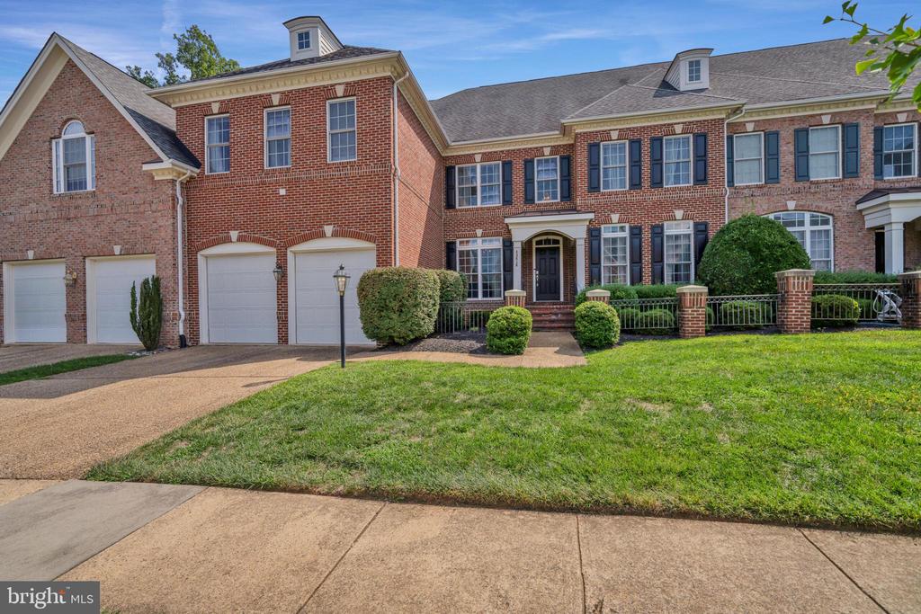 Large red-brick house with multiple garage doors, dark shutters, landscaped front lawn, and shrubs.