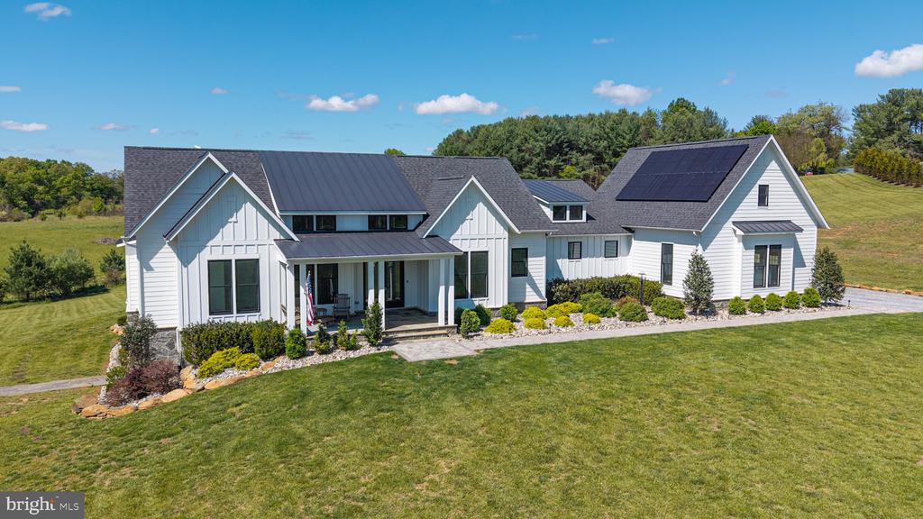 White farmhouse-style home with a covered porch, solar panels, landscaped beds, and a broad lawn.