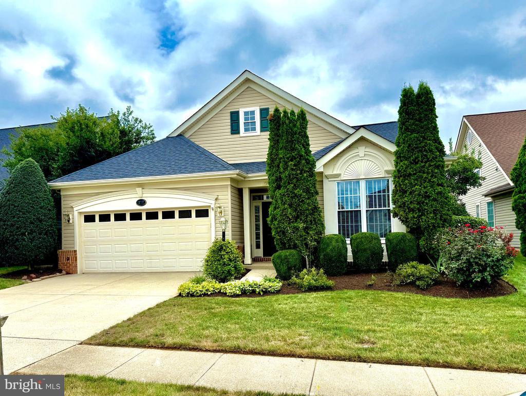 Beige home with a two-car garage, tall evergreens, landscaped shrubs, and a front lawn.