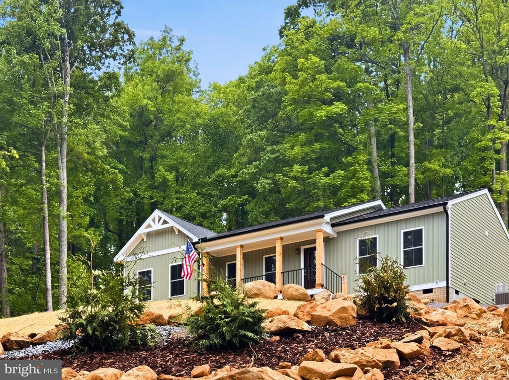Light gray home with a covered porch, American flag, and rocky landscaping in a wooded setting.