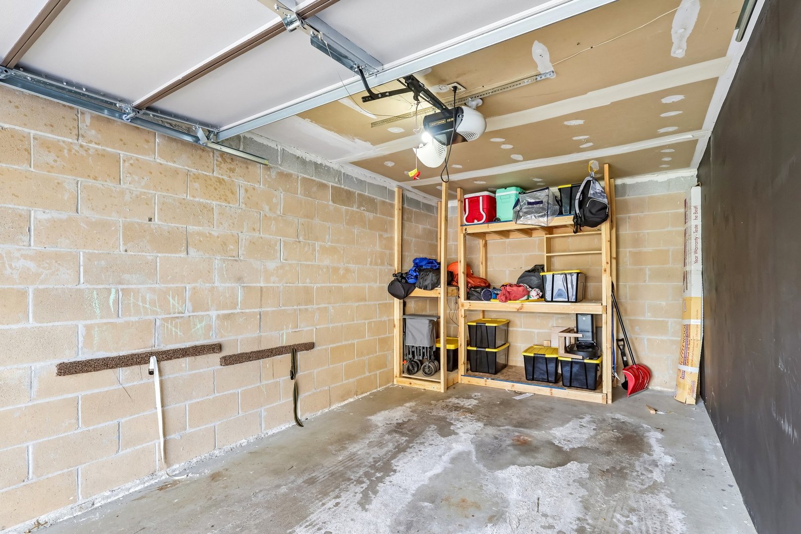 Garage with tan block walls, overhead door, and wooden storage shelves filled with bins and gear.