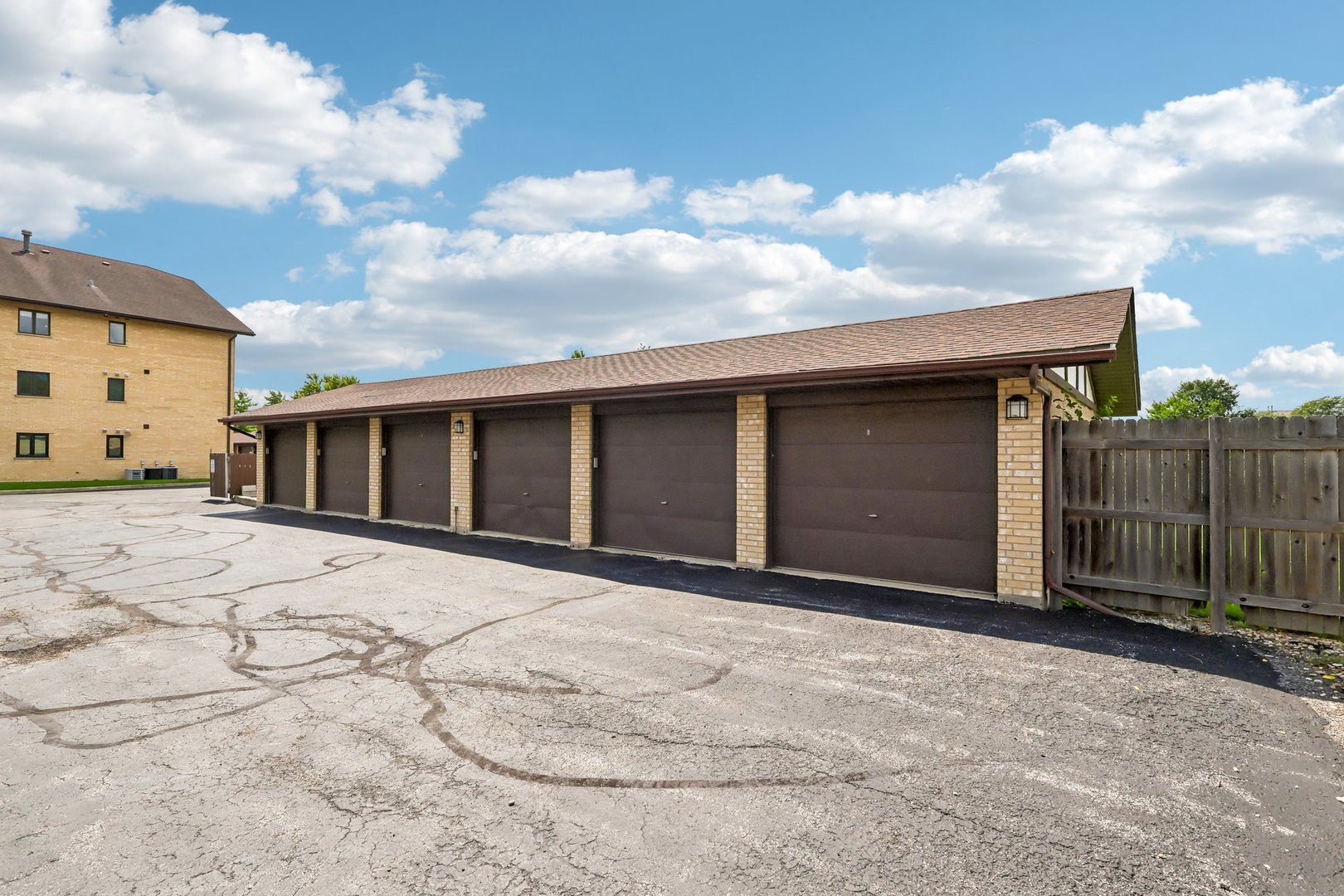 Attached brick garages with dark brown doors beside a cracked parking area and wooden fence.