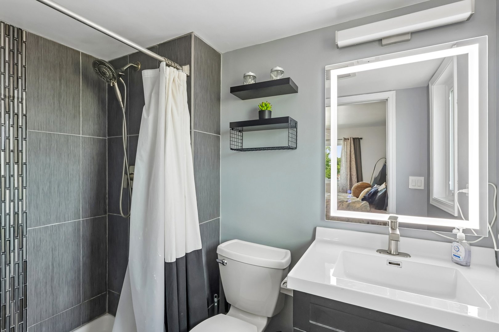Modern bathroom with a dark tiled shower, white fixtures, and an illuminated vanity mirror.