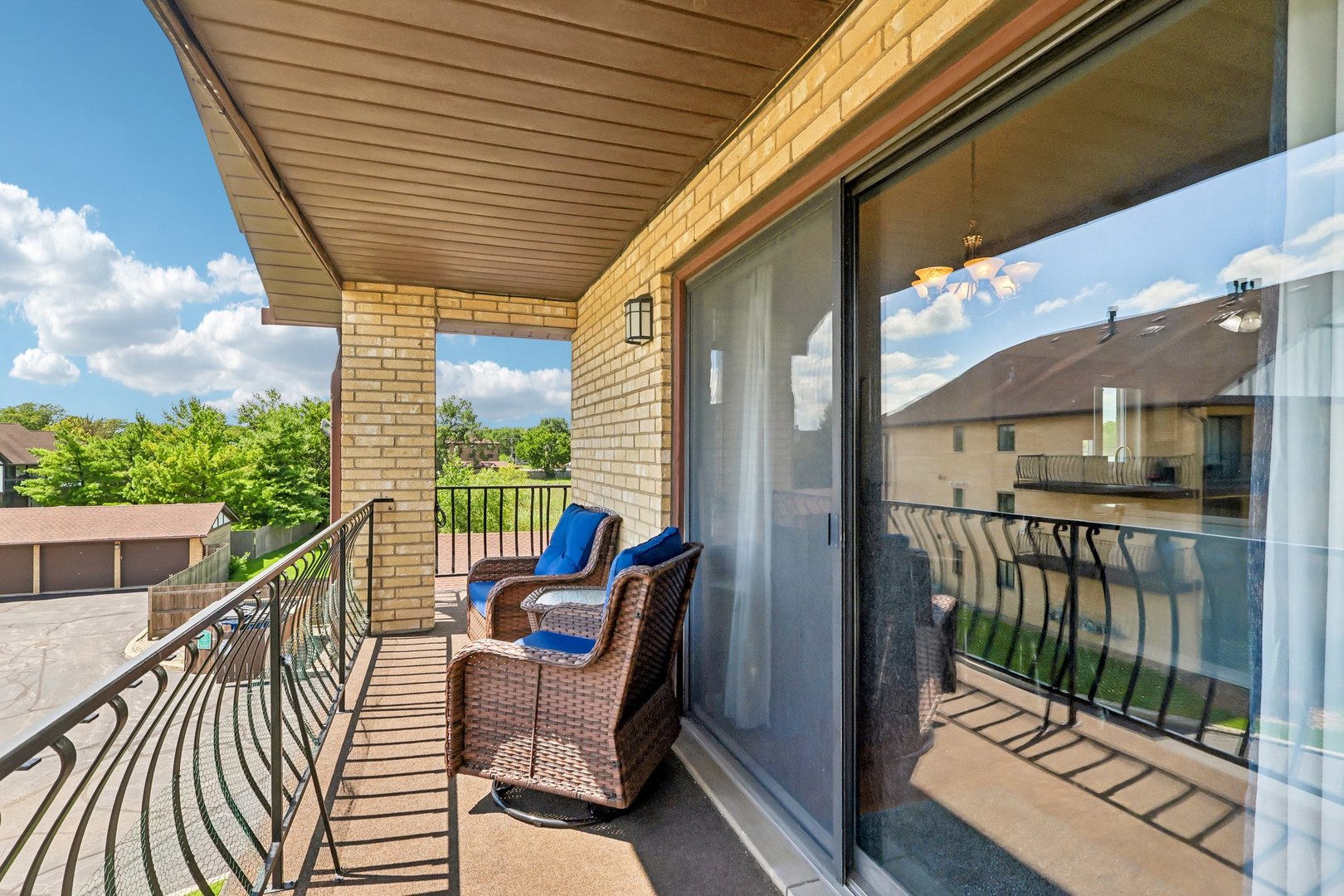 Covered balcony with wicker chairs beside sliding glass doors and a metal railing.
