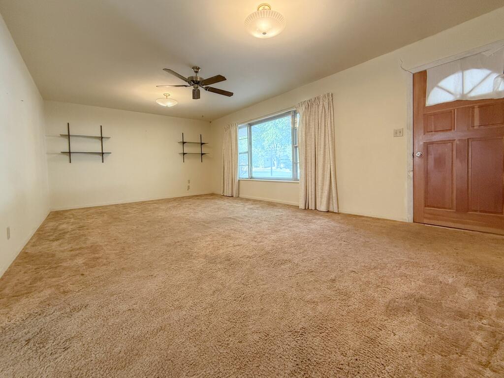 Vacant living room with beige carpet, wall shelves, a ceiling fan, and a large curtained window.