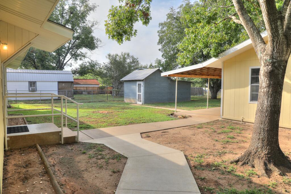 Yard with a covered patio, concrete walkway, metal outbuildings, and a large shade tree.