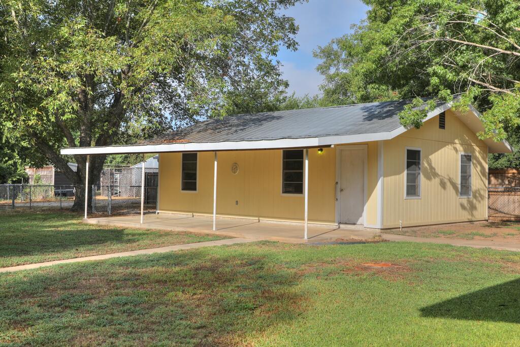Single-story yellow house with a dark metal roof and covered front porch beneath mature trees.