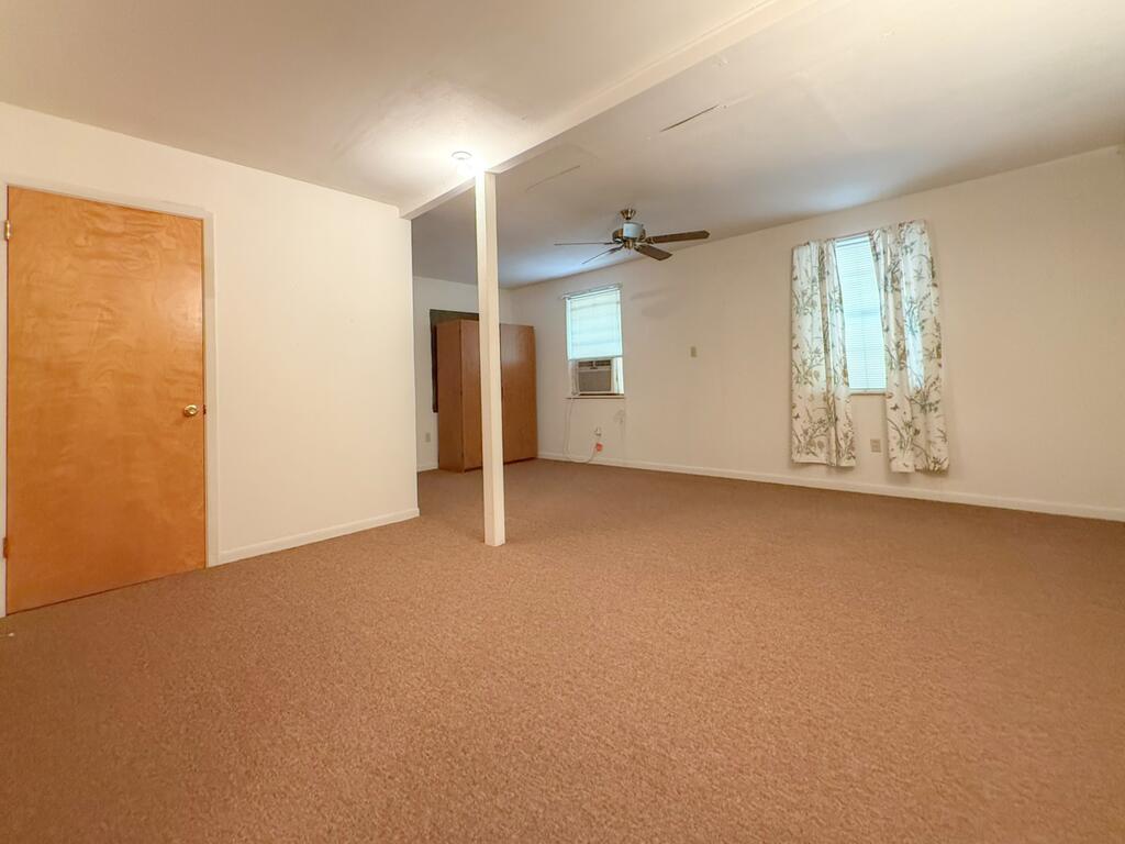 Vacant carpeted room with a ceiling fan, window air conditioner, and patterned curtains.