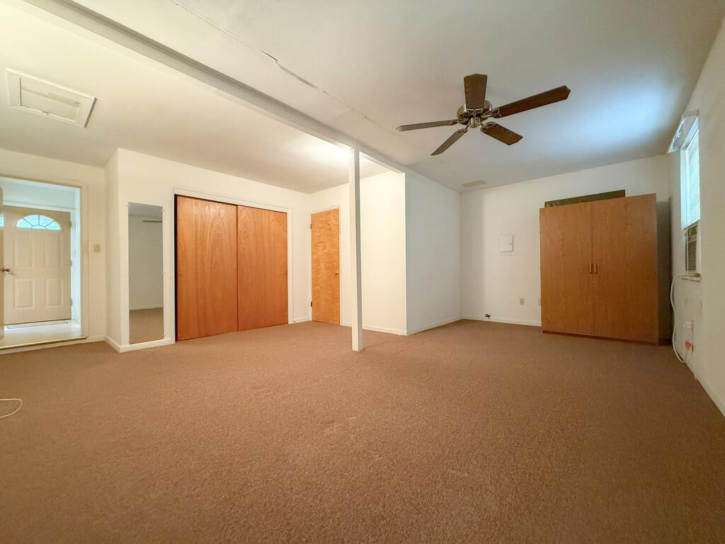 Empty bedroom with beige carpet, wooden closets, ceiling fan, and window air conditioner.