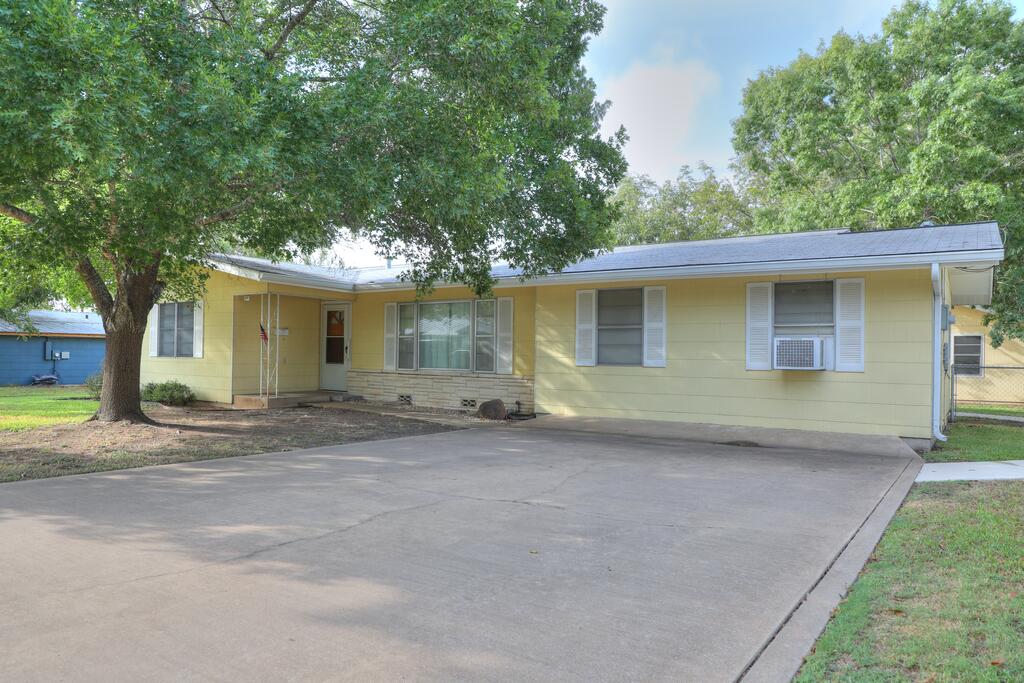 Single-story yellow house with white shutters, a wide concrete driveway, and a large shade tree.