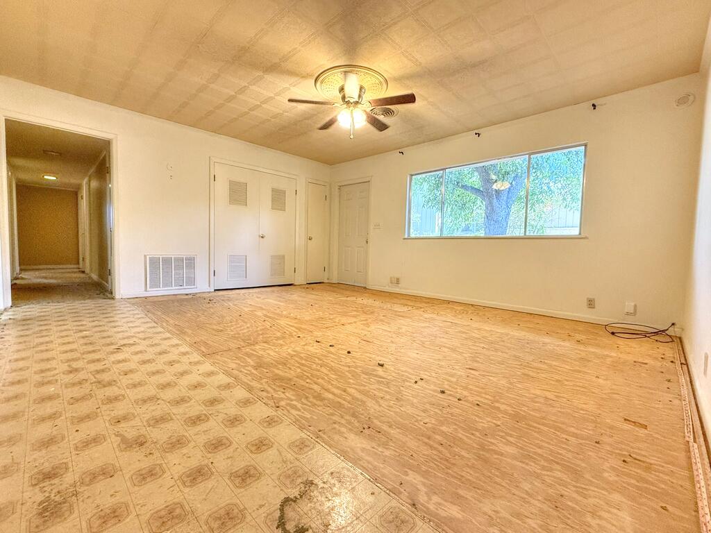 Vacant living room with a ceiling fan, wide window, and exposed plywood flooring.