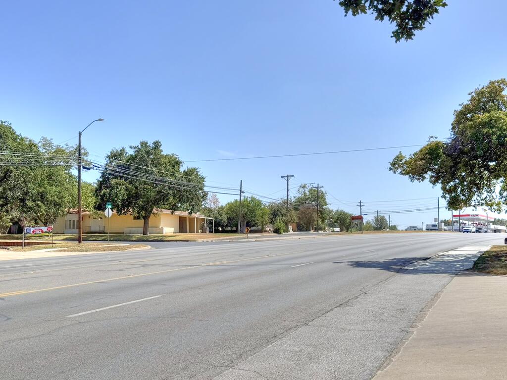 Single-story pale yellow building across a broad road, shaded by mature trees.