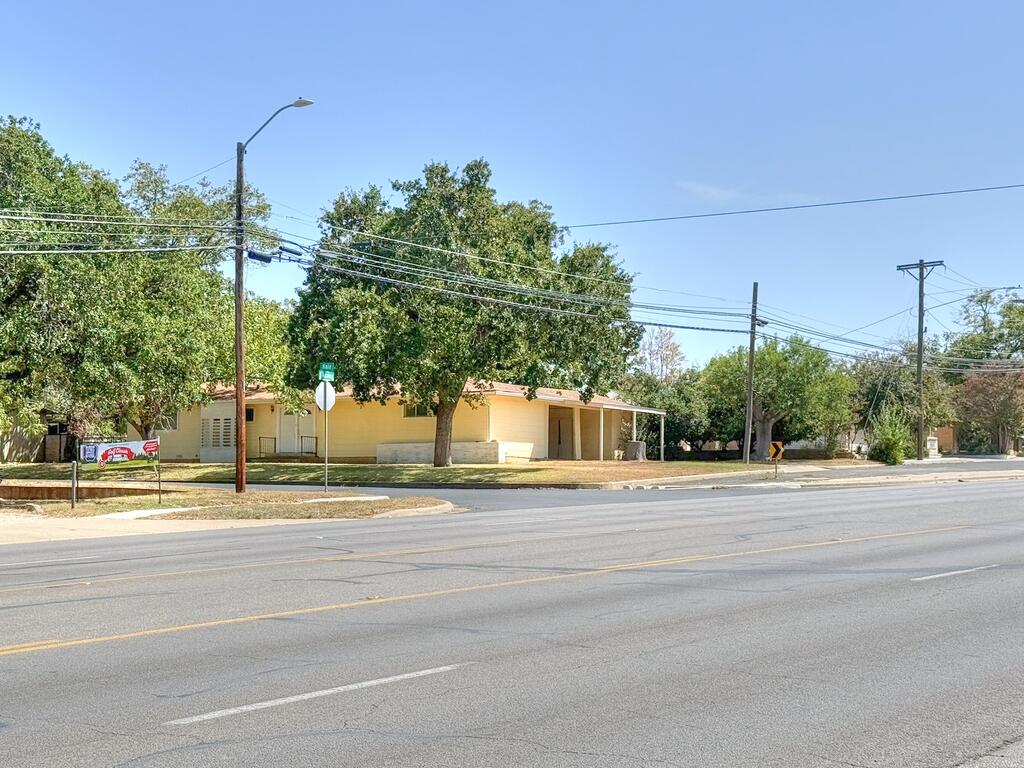 Single-story yellow building shaded by mature trees beside a broad street.