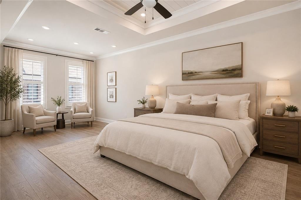 Staged bedroom with a large upholstered bed, tray ceiling, and two armchairs beside tall windows.