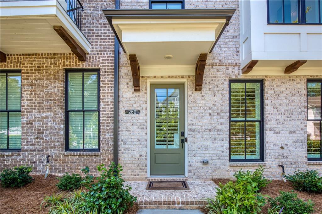 Brick home entrance with a sage-green door, covered porch, black-framed windows, and landscaped beds.