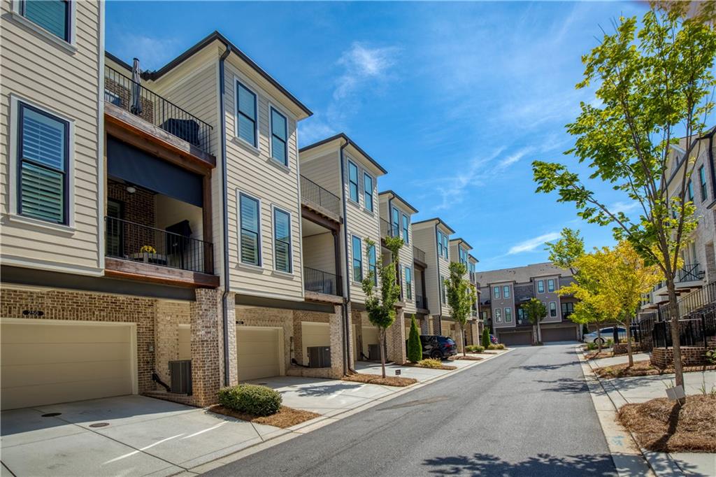 Attached townhomes line a sunny street, with brick lower levels, garage doors, and balconies.