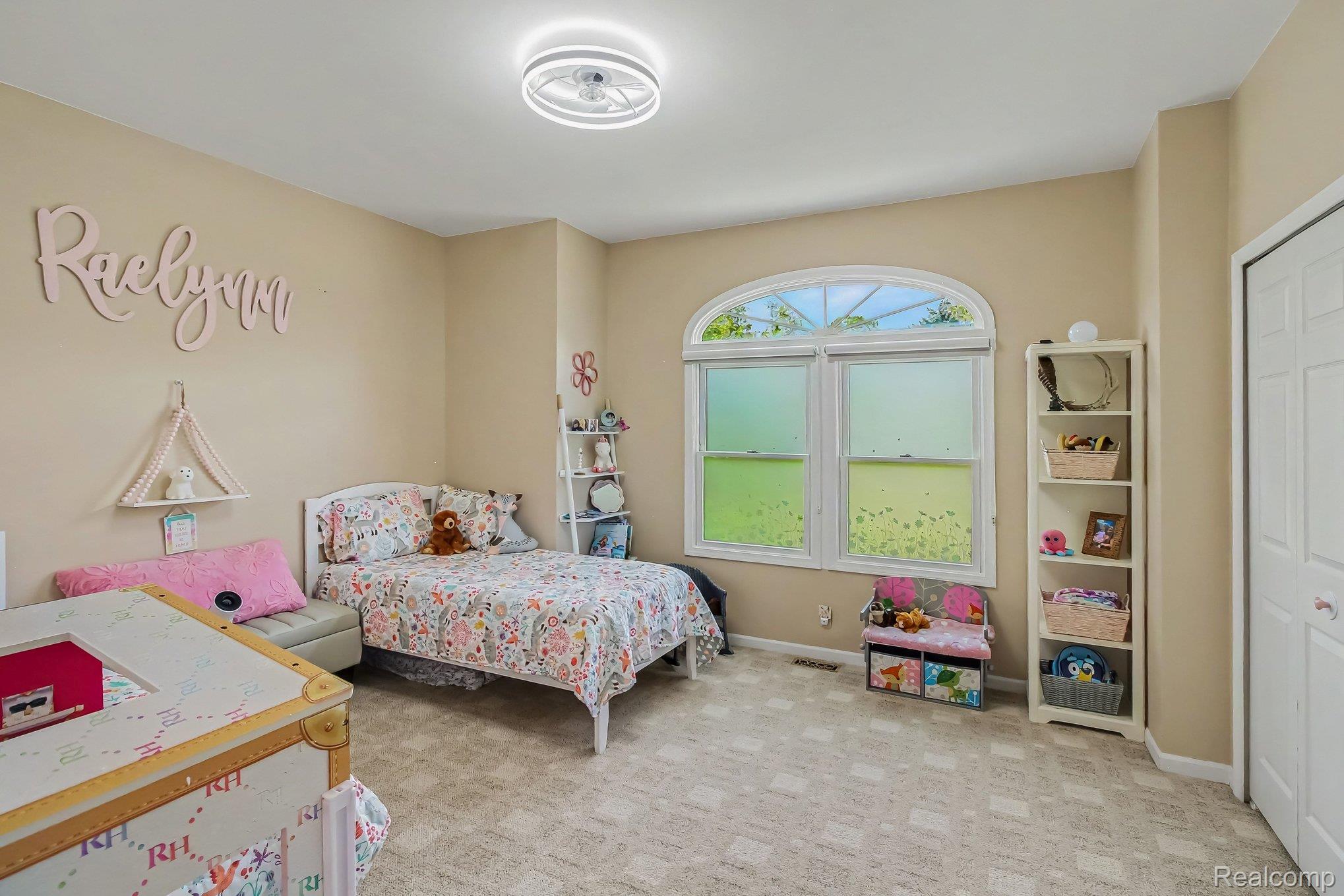 Furnished bedroom with floral bedding, an arched window, and a wall sign reading “Raelyn.”