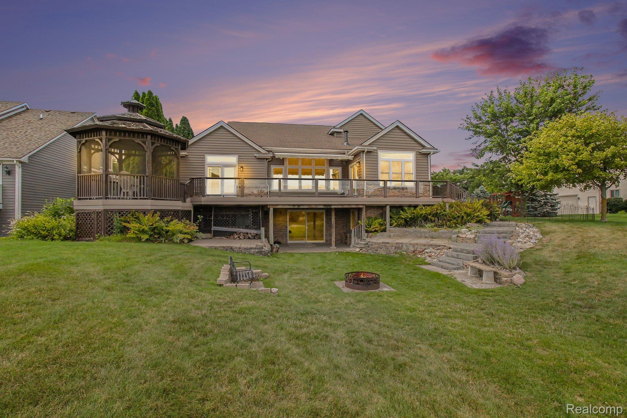 House rear with raised deck, screened gazebo, landscaped lawn, and fire pit at dusk.