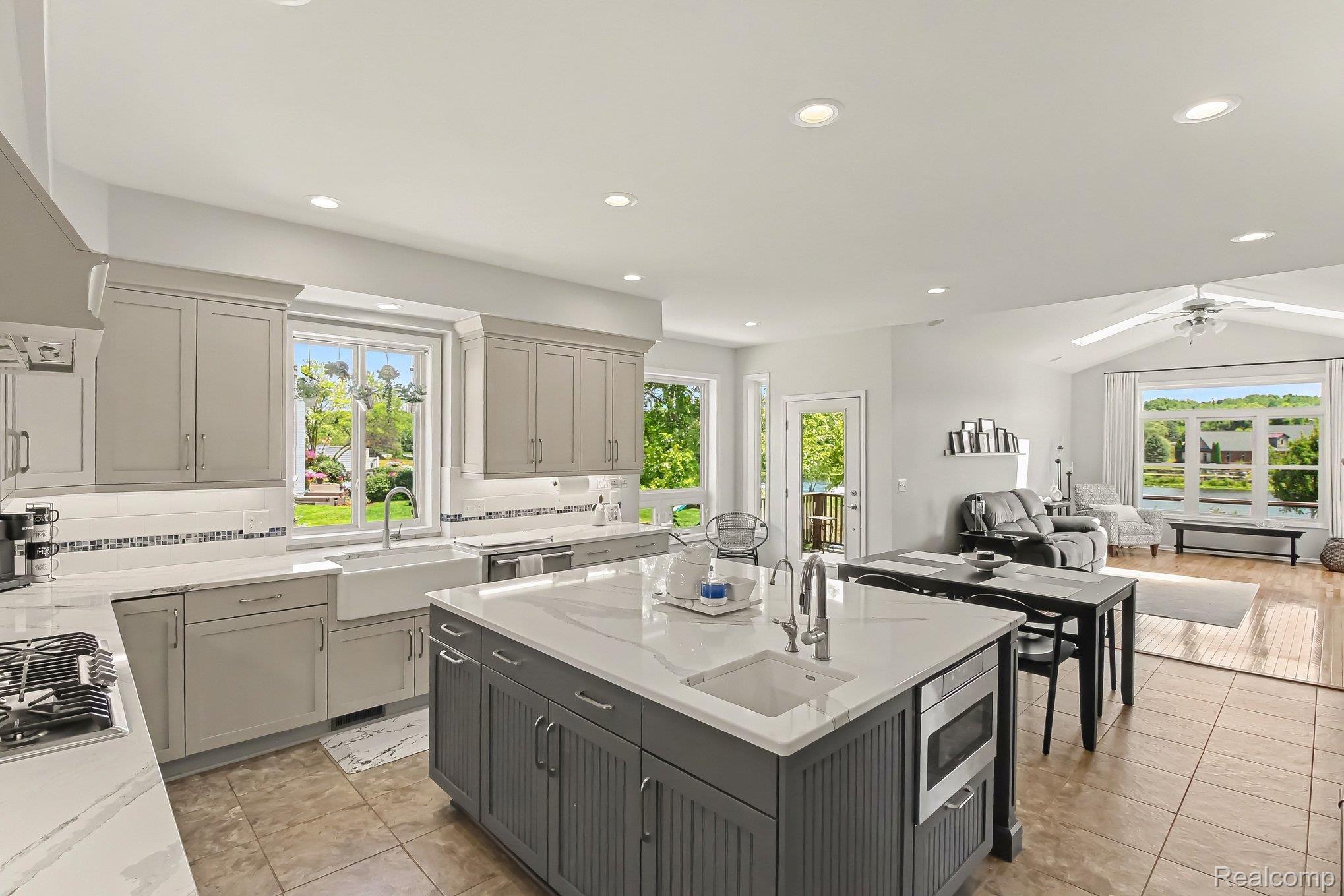 Kitchen with a large gray island and sink, opening to a living area with wide windows overlooking water.